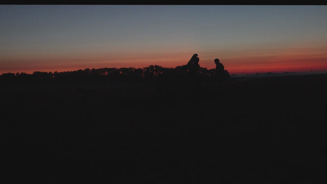 Silhouetted waterfowl hunters and a dog at the edge of a wetland beneath an orange sunset.